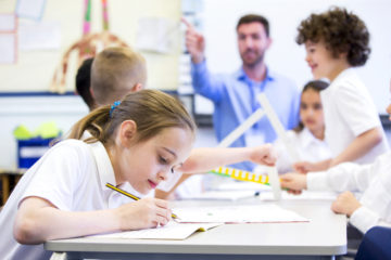 White female pupil writing with children and male teacher in background